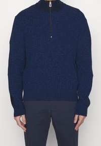 Blauer Strickpullover mit hohem Kragen, halbem Reißverschluss und langen Ärmeln, die mit strukturiertem Stoff und gerippten Bündchen versehen sind.