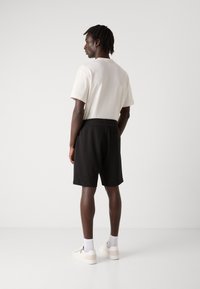 Pantaloni corti neri intrecciati con cintura elastica, abbinati a una camicia a maniche corte di colore chiaro. Sneakers bianche con accenti grigi e calzini alla caviglia.