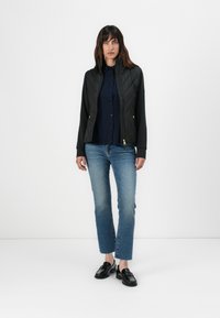 Zwarte gewatteerde jas met hoge kraag, ritsluiting, gecombineerd met een marineblauw overhemd met knopen en blauwe recht-pasvormige spijkerbroek. Zwarte instappers maken de outfit af.