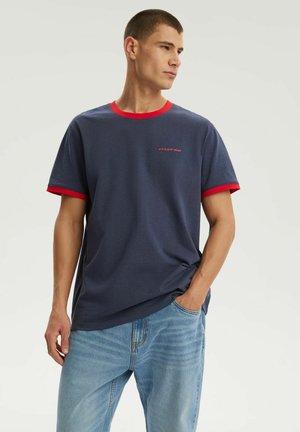 Mężczyzna ubrany w granatowy T-shirt z czerwonym kołnierzykiem i krawędziami rękawów, jasnoniebieskie dżinsy, patrzący na bok z jedną ręką w kieszeni.