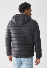 Hombre de espaldas vestido con una chaqueta acolchada con capucha negra y jeans de color azul claro frente a un fondo claro y liso.