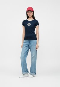 Μπλούζα ναυτικού με λευκές λεπτομέρειες, γραφικό λογότυπο στο στήθος, συνδυασμένη με ανοιχτό μπλε φαρδύ τζιν και λευκά αθλητικά παπούτσια, ολοκληρωμένο με κόκκινο καπέλο.