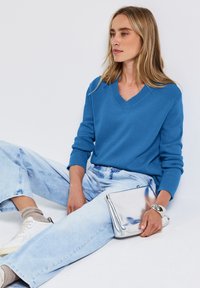 Pull bleu en V avec des poignets côtelés, associé à un jean large en denim bleu clair. Tenant une pochette argentée brillante. Design minimaliste.