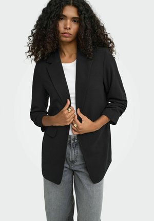 Kvinde med krøllet hår iført en sort blazer over en hvid top og grå jeans, der holder blazeren foran med begge hænder.