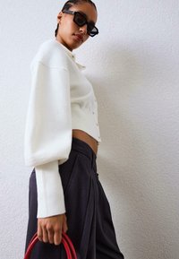 Biely pletený kardigán s nadmernými rukávmi, zapínaním na gombíky, v kombinácii s tmavými nohavicami a červenou kabelkou. Hladká textúra a moderný tvar.