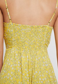 Gul blomsterklänning med smockad rygg, med tunna justerbara axelband, en rynkad textur och ett mönster av små vita blommor.
