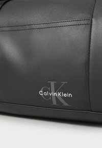 Schwarze Tasche aus synthetischem Leder mit glatter Textur, ausgestattet mit geprägtem "Calvin Klein"-Logo und passender Naht. Verfügt über ein minimales Design.