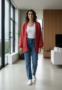 Czerwony, puszysty kardigan na białym topie, w połączeniu z niebieskimi dżinsami i białymi trampkami. Stoi w nowoczesnym wnętrzu z dużymi oknami.