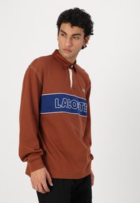 Hnedé polokošele s dlhým rukávom, ktoré majú biely golier a modrý horizontálny pruh s nápisom "LACOSTE" v bielom písmone. Vyrobené z bavlnenej tkaniny.