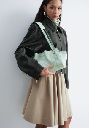 Moteris, apsirengusi juoda odine striuke ir smėlio spalvos sijonu, laiko šviesiai žalią odinę peties rankinę, stovėdama priešais neryškų foną.