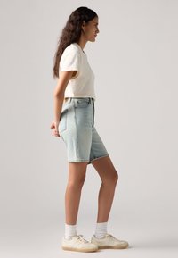 Κομμένο κρεμ T-shirt συνδυασμένο με ανοιχτόχρωμες τζιν σορτς. Το μοντέλο φοράει λευκά αθλητικά παπούτσια και κάλτσες μέχρι τον αστράγαλο, δείχνοντας μια χαλαρή εμφάνιση.