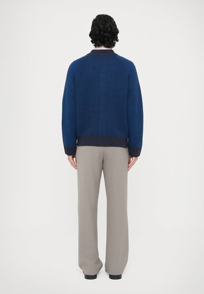 Blauer Strickpullover mit strukturiertem Muster, ausgestattet mit einem dunklen, gerippten Bund und Bündchen, kombiniert mit hellgrauen Hosen.