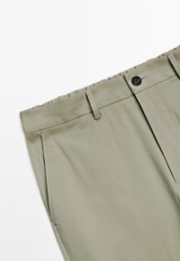 Pantaloni beige chiaro con chiusura a bottone, passanti per cintura e tasche laterali su sfondo bianco, con focus sull'area della vita.