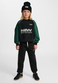 Czarna bluza z zielonymi rękawami, z logo, w połączeniu z czarnymi spodniami dresowymi z bocznymi paskami. Czapka beanie i trampki dopełniają strój.