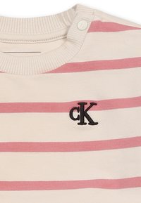 T-shirt a righe con strisce orizzontali crema e rosa, dotata di scollo a girocollo a coste e un logo "cK" ricamato in nero sul petto. Due bottoni a pressione sul collo.