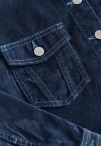 Granatowy materiał sztruksowy z przednią kieszenią na piersi i zapięciem na srebrny zatrzask. Charakteryzuje się kontrastującym jasnoniebieskim przeszyciem. Teksturowana powierzchnia.