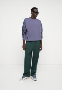 Purple sweatshirt with striped sleeves, paired with dark green wide-leg pants and light blue sneakers. Model wearing dark sunglasses.