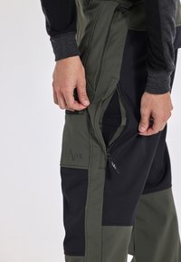 Technische broeken in een olijfgroen en zwart kleurenschema, met ritsen aan de zijkanten, duurzaam materiaal en een soepele textuur.