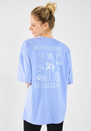 Tricou oversized din bumbac bleu deschis, cu grafice albe pe spate care prezintă icoane legate de călătorie și fraza "Aventura te cheamă."