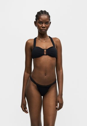 Kobieta z warkoczami, ubrana w czarne dwuczęściowe bikini z topem na szyję i minimalną biżuterią na jednolitym jasnym tle.