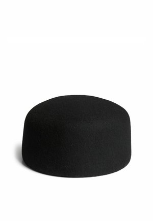 Jeune femme souriante, portant une veste en fausse fourrure grise et un chapeau noir, les bras croisés devant un fond blanc uni.