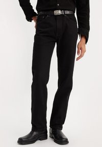 Personne portant un jean noir ajusté avec une ceinture noire, une chemise noire à boutons et des bottines en cuir noir sur fond blanc.