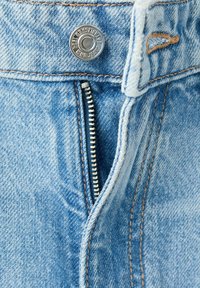 Gros plan sur un jean en denim bleu clair avec une fermeture éclair partiellement ouverte et un bouton argenté marqué « KING JEANS Est. 1964 ». Coutures marron visibles.