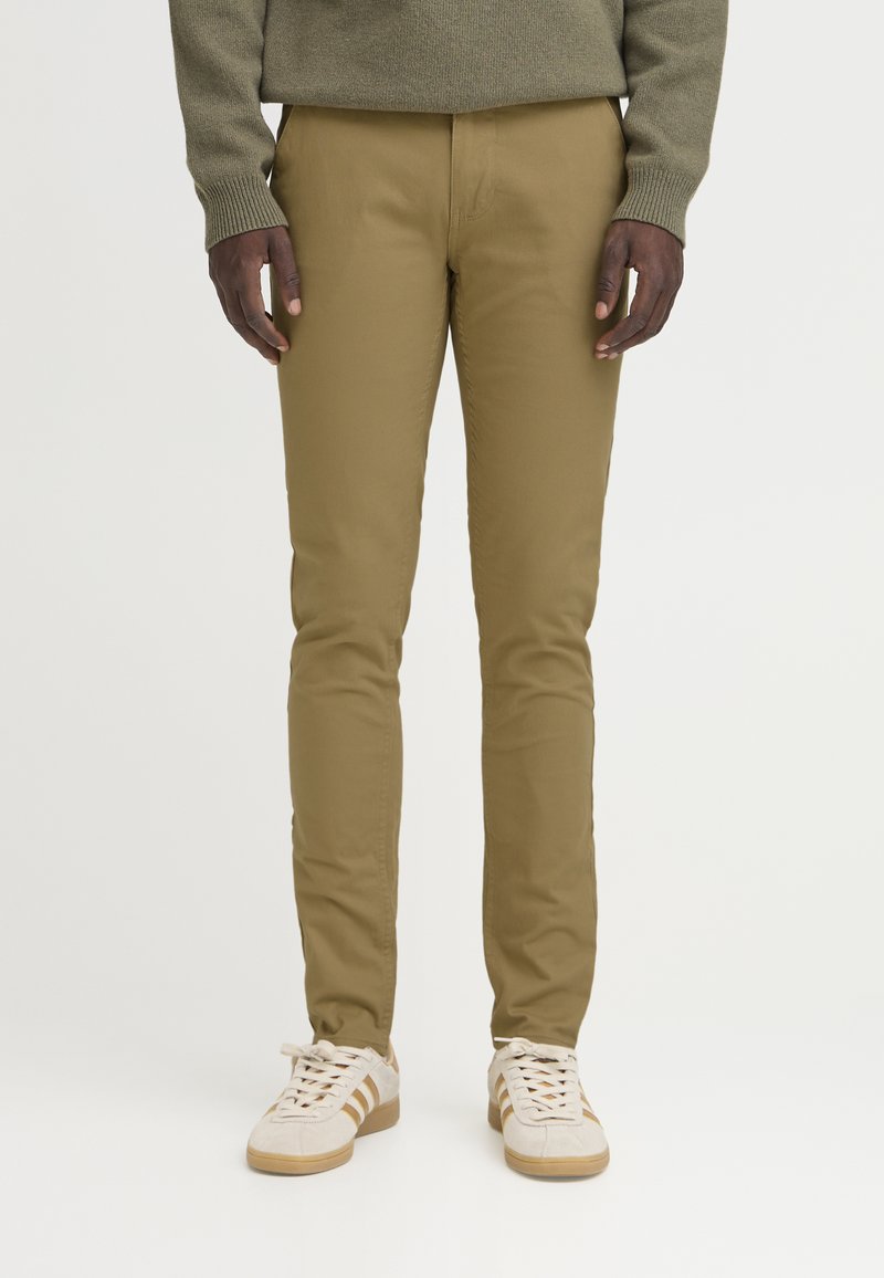 Muž nosící slim-fit khaki kalhoty, olivově zelený svetr a bílé tenisky s béžovými pruhy, stojící před jednobarevným pozadím.