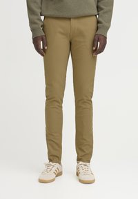 Pantalones caqui con un corte ajustado y una textura suave, combinados con zapatillas deportivas beige que presentan tres rayas. Diseño simple y funcional.