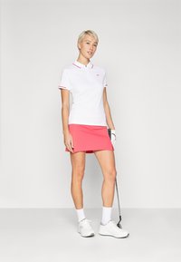 Chemise polo blanche avec des accents rouges, associée à une jupe rose. Le modèle tient un club de golf, portant des chaussures et des chaussettes blanches. Design simple.