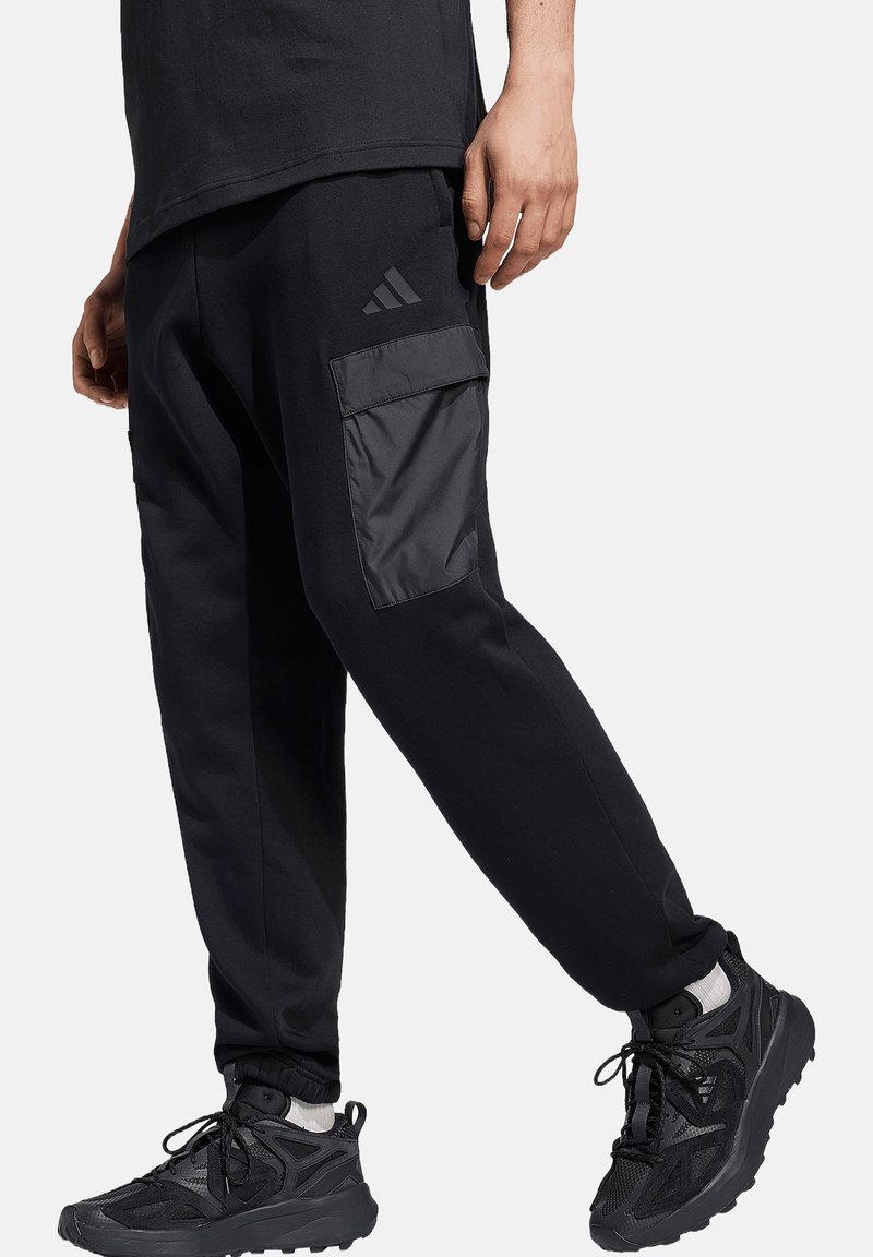 Schwarze Jogginghosen mit einer glänzenden schwarzen Seitentasche aus weichem Material, ausgestattet mit gerippten Bündchen und getragen mit schwarzen Sportschuhen.