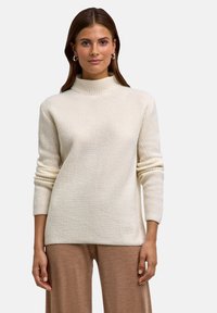 Maglione a coste di colore bianco sporco con collo alto e maniche lunghe. Tessuto morbido, vestibilità comoda, abbinato a pantaloni beige a gamba larga.