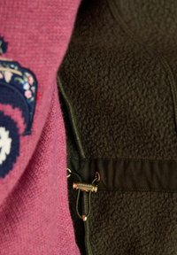 Zbliżenie różowego dzianinowego materiału obok ciemnobrązowego polaru z czarnym sznurkiem i małym metalowym zapięciem oznaczonym "Joules".