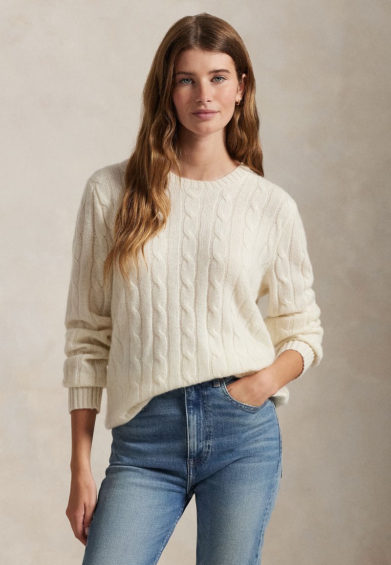 Suéter creme de malha cable-knit com gola redonda e mangas dobradas, combinado com jeans de algodão azul claro. A textura é macia com padrões em relevo.