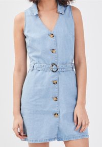 Robe en denim bleu clair sans manches avec des boutons sur le devant et une ceinture assortie avec une boucle en métal ronde, portée par une personne aux cheveux bouclés.