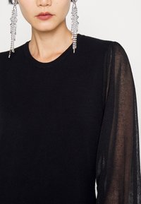 Femme portant un pull noir avec manches longues transparentes et longues boucles d'oreilles pendantes argentées, couleur de lèvres neutre et visage recadré.