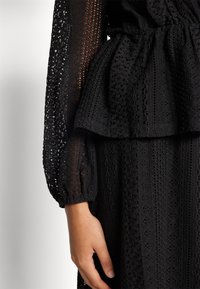 Abito nero in tessuto texture con design peplum, maniche lunghe con polsini arricciati e intricate decorazioni di pizzo sul corpetto e sulla gonna.