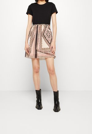 Person wearing a black short-sleeve top, patterned beige and pink pleated skirt, and black ankle boots standing against a white background.