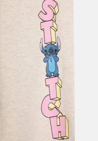 Krämfärgad t-shirt med en blå tecknad karaktär med stora öron, tillsammans med fet rosa och gula 3D-bokstäver som stavas "STITCH."
