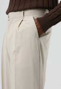 Beige broek met plooien aan de voorkant, zijzakken en een gladde textuur, gecombineerd met een bruine geribde gebreide trui.