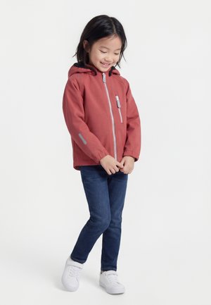 Νεαρή κοπέλα που χαμογελά, φοράει μπουφάν με κουκούλα σε σκουροκόκκινο χρώμα, σκούρο μπλε τζιν και λευκά αθλητικά παπούτσια, στέκεται με τα χέρια ενωμένα.