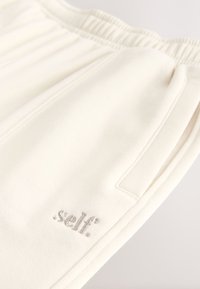 Kreminės spalvos sportinės kelnės iš minkštos medžiagos, su elastinga juosmens juosta ir šoninėmis kišenėmis. Prekės ženklo logotipas išsiuvinėtas pilka spalva.