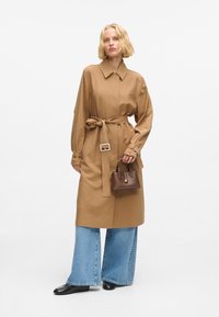 Cappotto trench beige con cintura, colletto, maniche larghe e tasche laterali, abbinato a jeans svasati blu chiaro e una piccola borsa marrone.