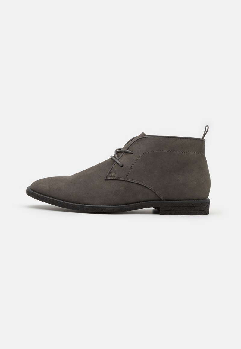 Bottine en daim gris foncé avec lacets, talon bas empilé et languette à l’arrière, présentée de profil sur un fond uni.