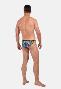Mann, der barfuß steht und sich abwendet, trägt bunte Schwimmshorts mit tropischem Muster vor einem weißen Hintergrund.