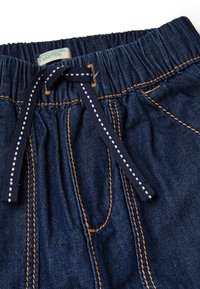 Tmavě modrý denimový pas s elastickým páskem a námořnickým šňůrkovým uzávěrem s bílými tečkami; kontrastní šití viditelné podél švů.