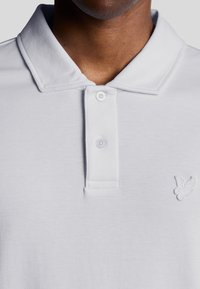 Mann trägt ein weißes Polohemd mit Kragen, zwei Knöpfen und einem kleinen gestickten Vogel-Logo auf der Brust.