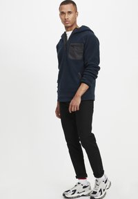 Marineblå fleece-pullover med en sort lynlåslomme på brystet, lange ærmer og en justerbar hætte, parret med sorte tapered bukser og sportssko.