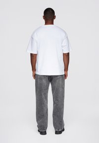 Weißes Baumwoll-T-Shirt mit lockerer Passform, von hinten gezeigt, kombiniert mit dunkelgrauen Denim-Jeans, die einen weiten Schnitt und Taschen aufweisen.