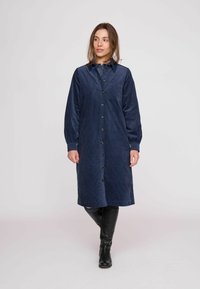 Námornícky modré šaty z kordurovej látky s predným zapínaním na gombíky, dlhými rukávmi, golierom a náprsným vreckom. Tkanina má rebrovanú textúru a siaha pod kolená.